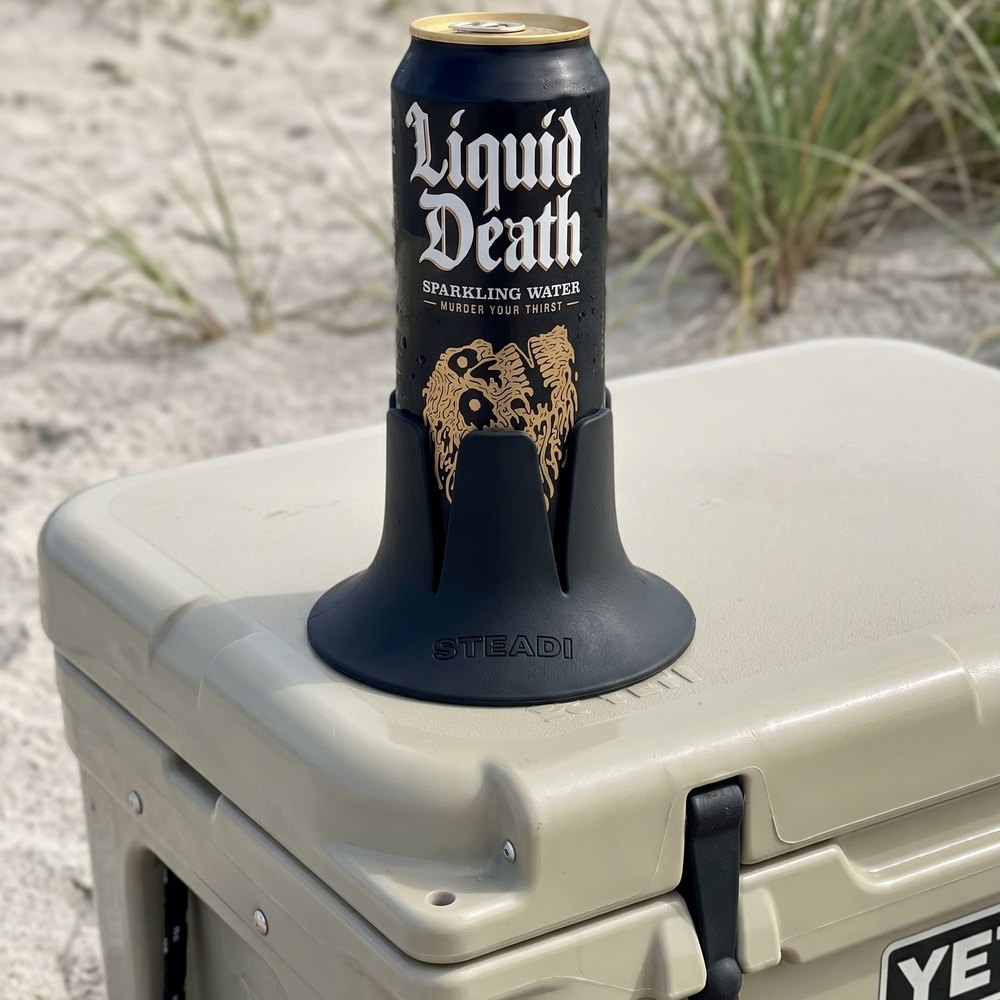 A tallboy can of sparkling water standing in a charcoal Steadi on the lid of a hard cooler, outdoors on open ground.