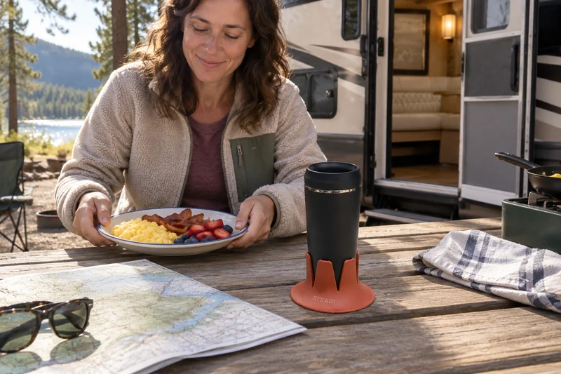 Adult making breakfast at campsite picnic table with open narrow-base reusable coffee cup in Steadi on level campsite picnic table with base fully supported between plank gaps. AI illustration.